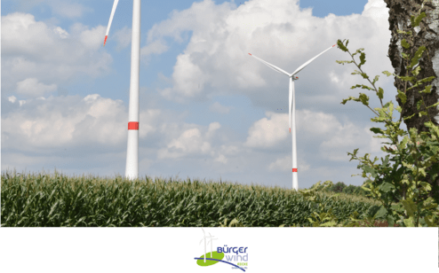 Zwei Windkraftanlagen hinter einer Maisfeldkante unter einem bewölkten Himmel. Logo von Bürgeer Wind unten.