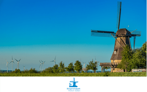 Windmühle neben Windkraftanlagen vor blauem Himmel, umgeben von Bäumen und Wiese im Bürgerwindpark Hollich Seilen.