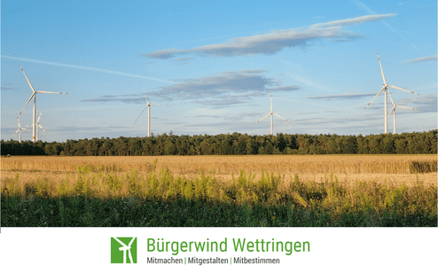 Windräder stehen neben einem Feld mit goldgelbem Getreide, umgeben von Bäumen und blauem Himmel.