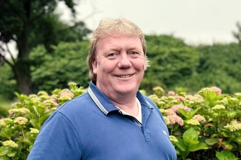 Lächelnder Mann in blauem Poloshirt steht vor einer Hecke mit blühenden Hortensien im Garten.