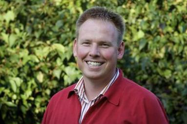 Lächelnder Mann mit kurzen Haaren, rot-weiß gestreiftem Hemd und rotem Pullover vor grüner Pflanzenwand.