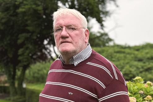 Älterer Mann in gestreiftem Pullover steht im Garten vor Sträuchern und dekorativen Schafsskulpturen.